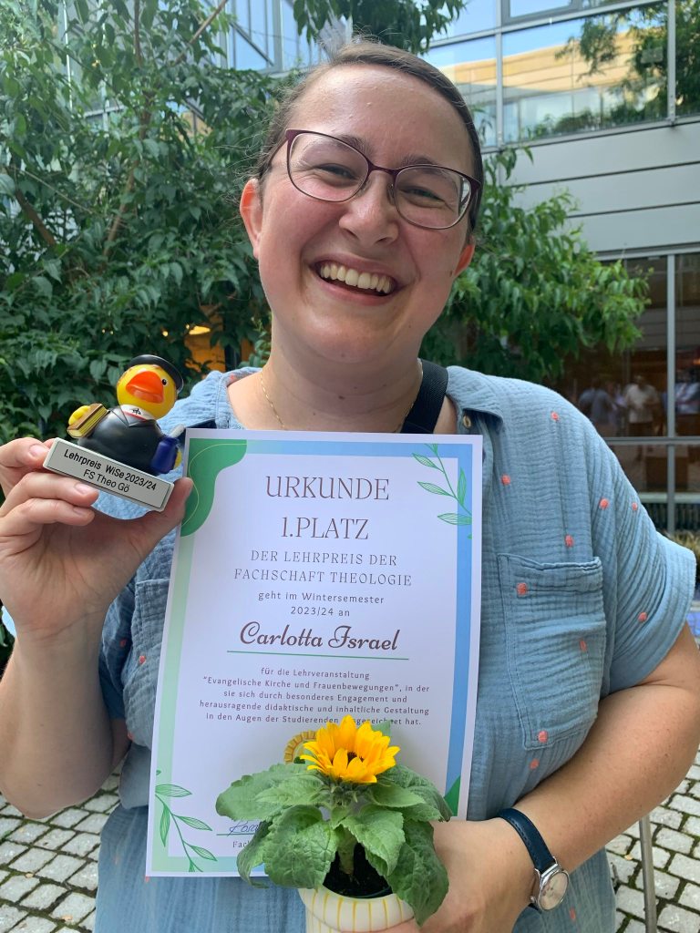 Auf dem Foto bin ich, Carlotta, zu sehen und halte eine Urkunde, deren Text ich im Fließtext zitiere, in der einen Hand und in der anderen Hand die Trophäe. Die Trophäe ist eine kleine Lutherente und hat als Unterschrift "Lehrpreis WiSe 2023/24 Fs Theo Gö", also Lehrpreis für das Wintersemester 2023/24 der Fachschaft Theologie Göttingen.